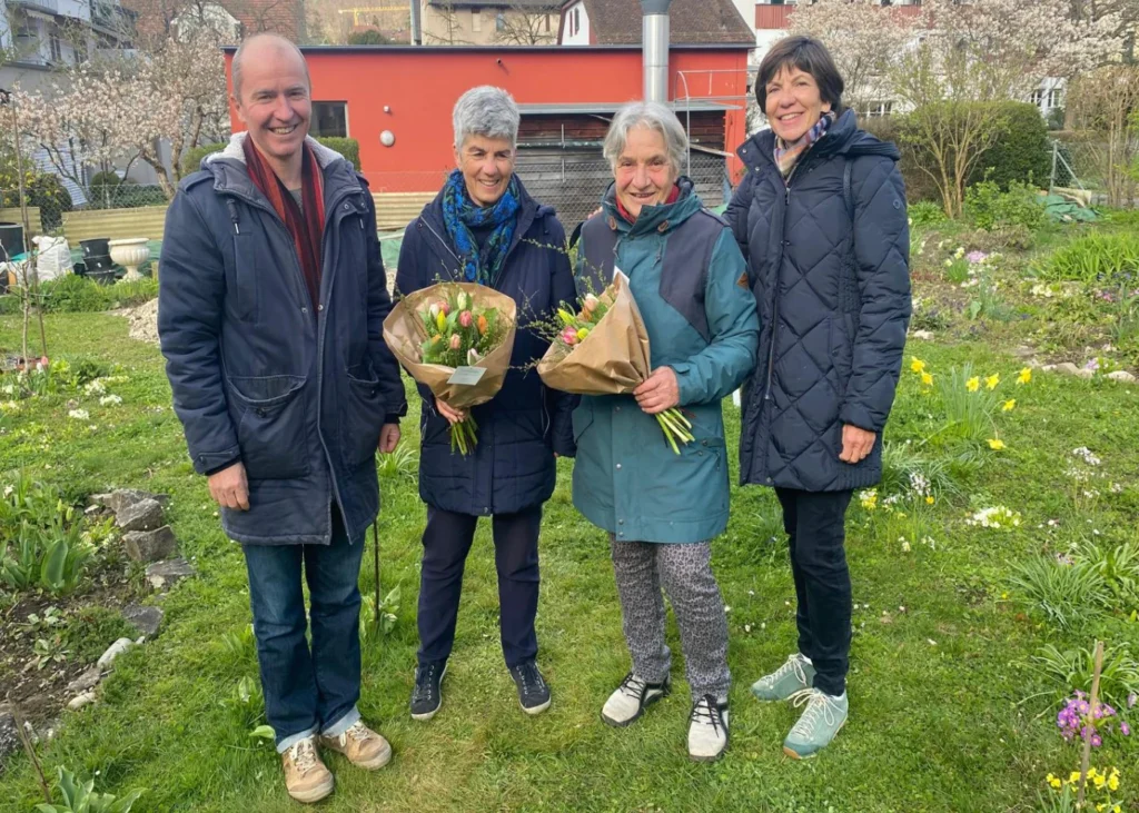 Four adults in a garden, two women holding bouquets wrapped in brown paper, smiling at the camera.