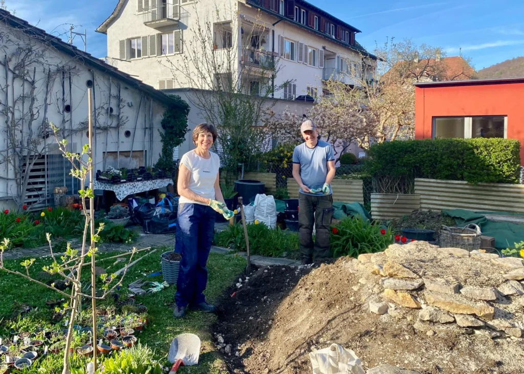 Two people gardening in a sunny backyard, pile of soil and rocks nearby, with flowering plants and tools around them, smiling at the camera.