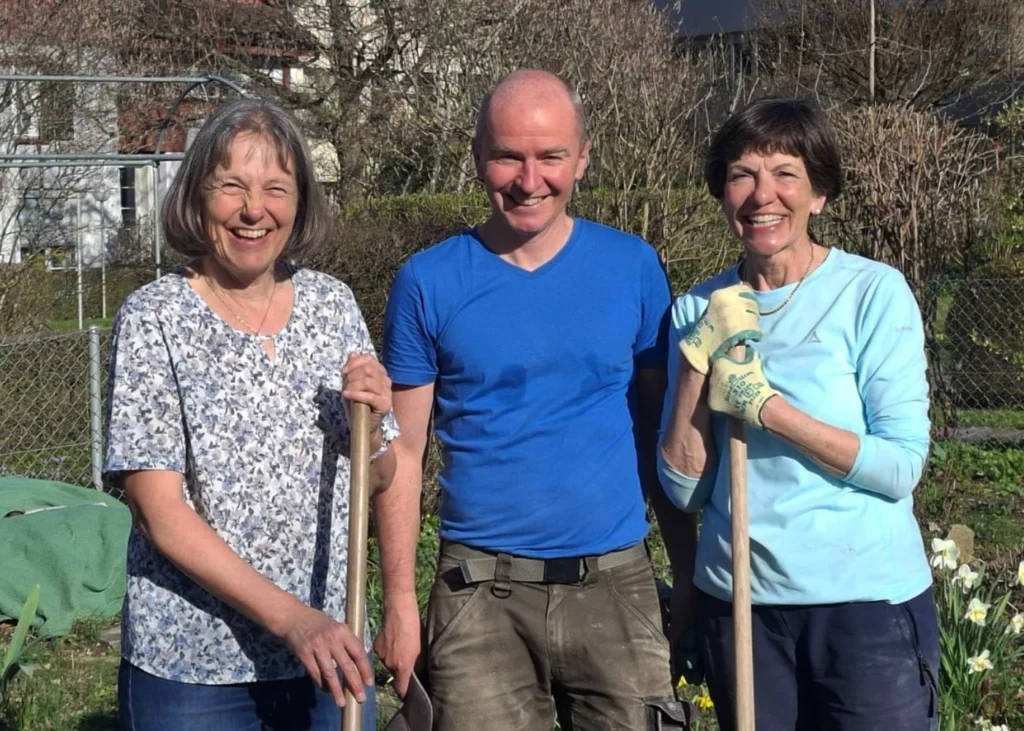 Three adults stand together in a garden, each holding a gardening tool and smiling at the camera.