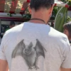 Man wearing a light gray T-shirt with a large bat illustration on the back, standing at an outdoor market under striped tents.