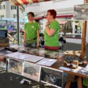 Two volunteers in green shirts stand behind a market stall table displaying framed wildlife photos, jars, and brochures, with a bat image hanging nearby.